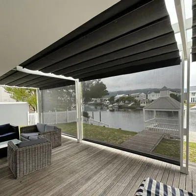 Clear outdoor blind enclosing a timber deck patio beside a river, with wicker seating