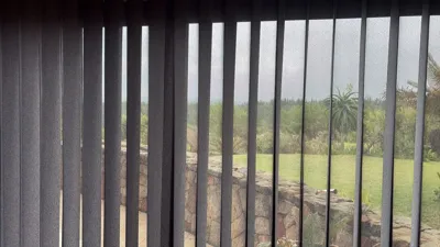 Dark grey vertical blinds at a window with a view of a stone garden wall, lawn and palm trees