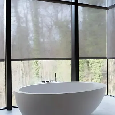 Grey sheer roller blinds on floor-to-ceiling windows beside a freestanding bath with forest view