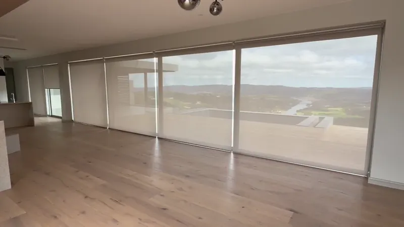 Cream sheer roller blinds raising together across a full-width glass wall on Somfy motors, revealing a valley and river view