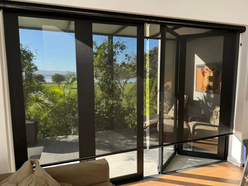 Charcoal sheer roller blinds by Custom Blinds Shutters & Awnings across full-width bifold doors, keeping the lagoon view while cutting glare
