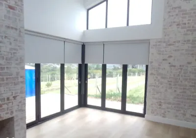 Light grey blockout roller blinds above corner bifold glass doors in a brick-walled lounge