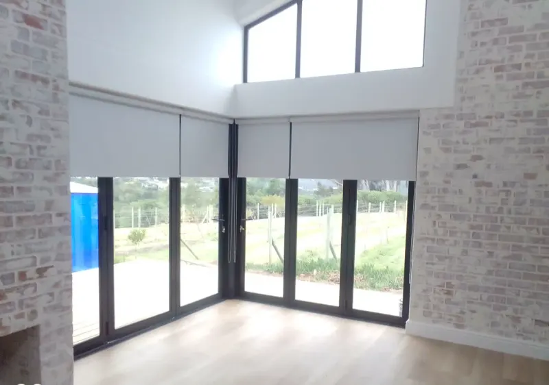 Riviera Mist blockout roller blinds by Custom Blinds Shutters & Awnings on corner bifold doors in a double-volume lounge with a brick feature wall
