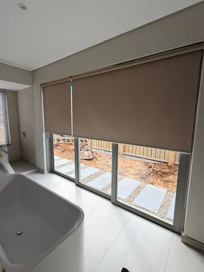 Taupe blockout roller blinds over bathroom sliding doors beside a freestanding bath