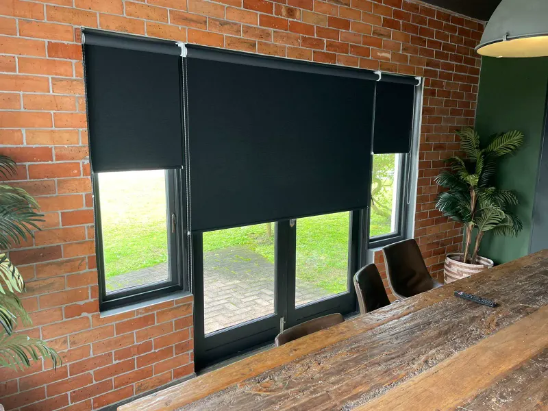 Three black blockout roller blinds at different drops over windows and doors in a face-brick dining room