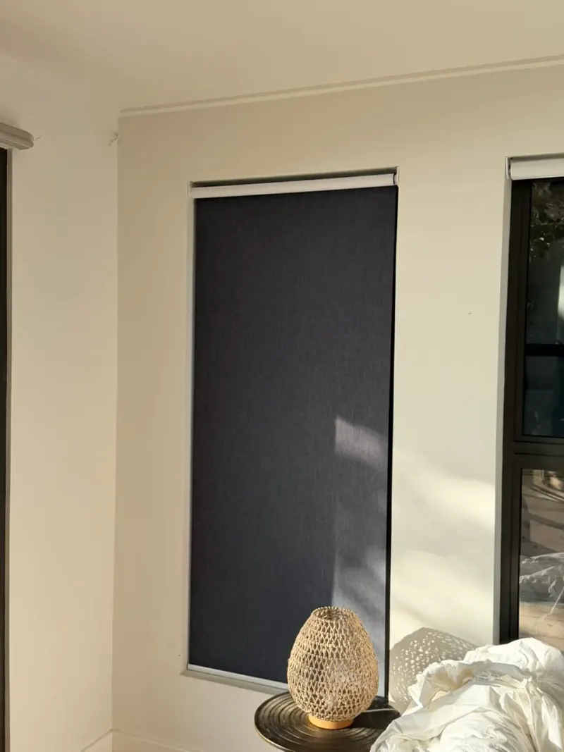 A charcoal blockout roller blind fully down on a bedroom window beside a bedside lamp