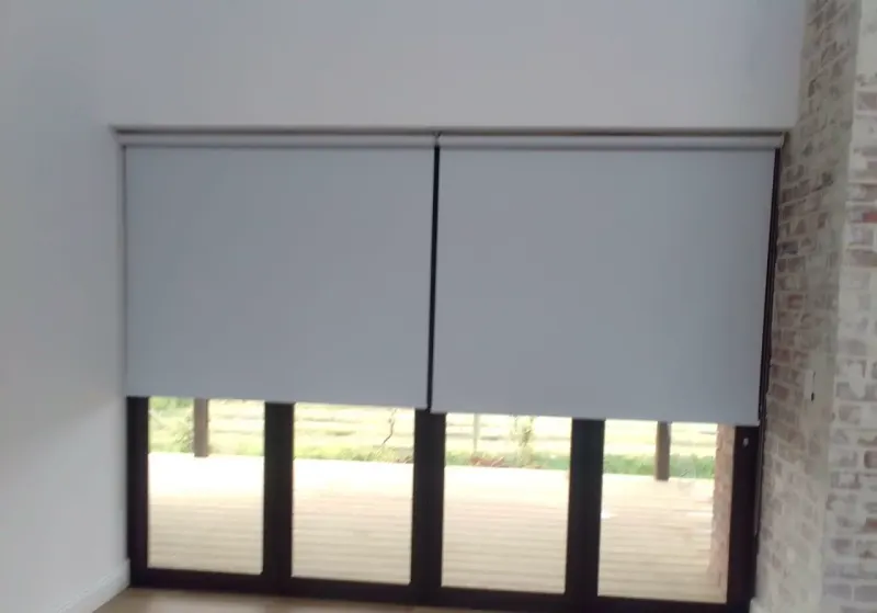 Two grey blockout roller blinds half down over stacking patio doors onto a timber deck