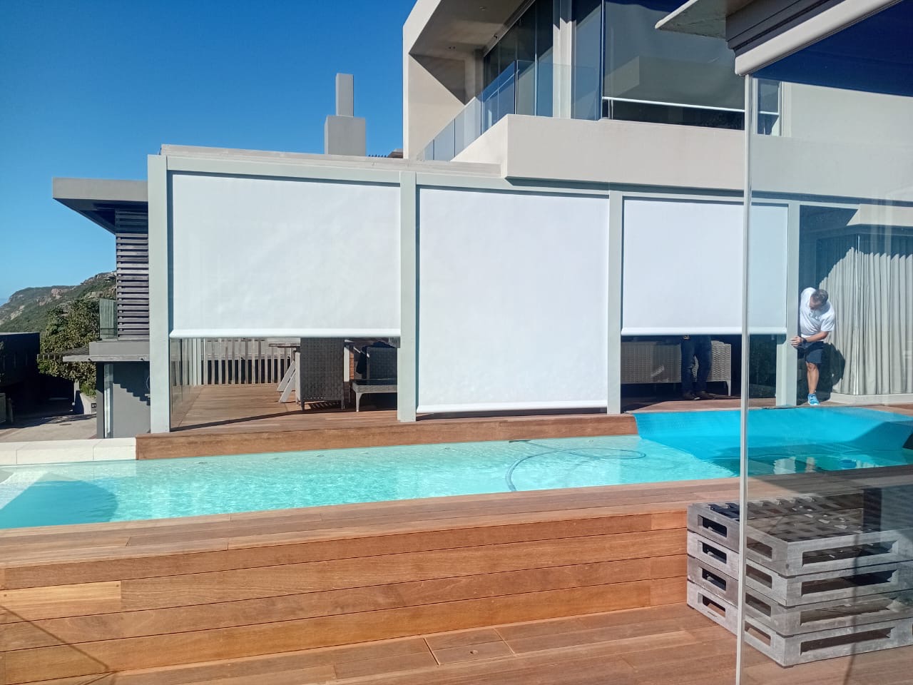 White outdoor screen blinds on pool-facing patio, mountain backdrop, coastal home
