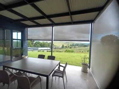 Outdoor screen blinds lowered on a covered patio with a dining table and green hills beyond