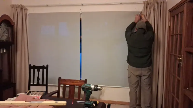 A sheer roller blind being fitted above a dining room window alongside existing curtains