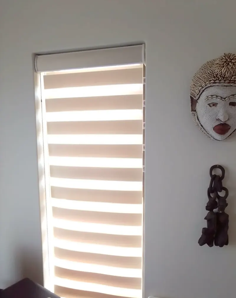 A cream zebra double roller blind filling a bedroom window with afternoon sun, a carved mask on the wall beside it